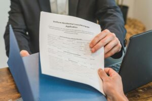 A man in a suit handing a Residential loan application to someone else with folders and a laptop on a desk between them